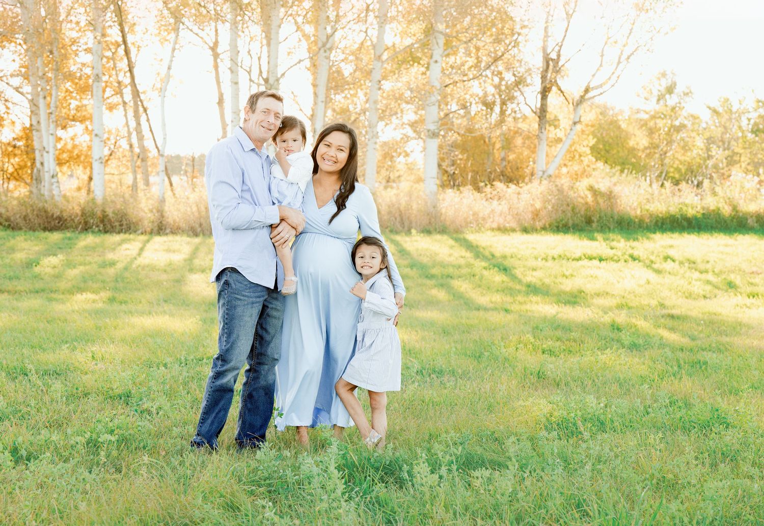 A family of four poses together in coordinating blue and white outfits in a sunlit field.