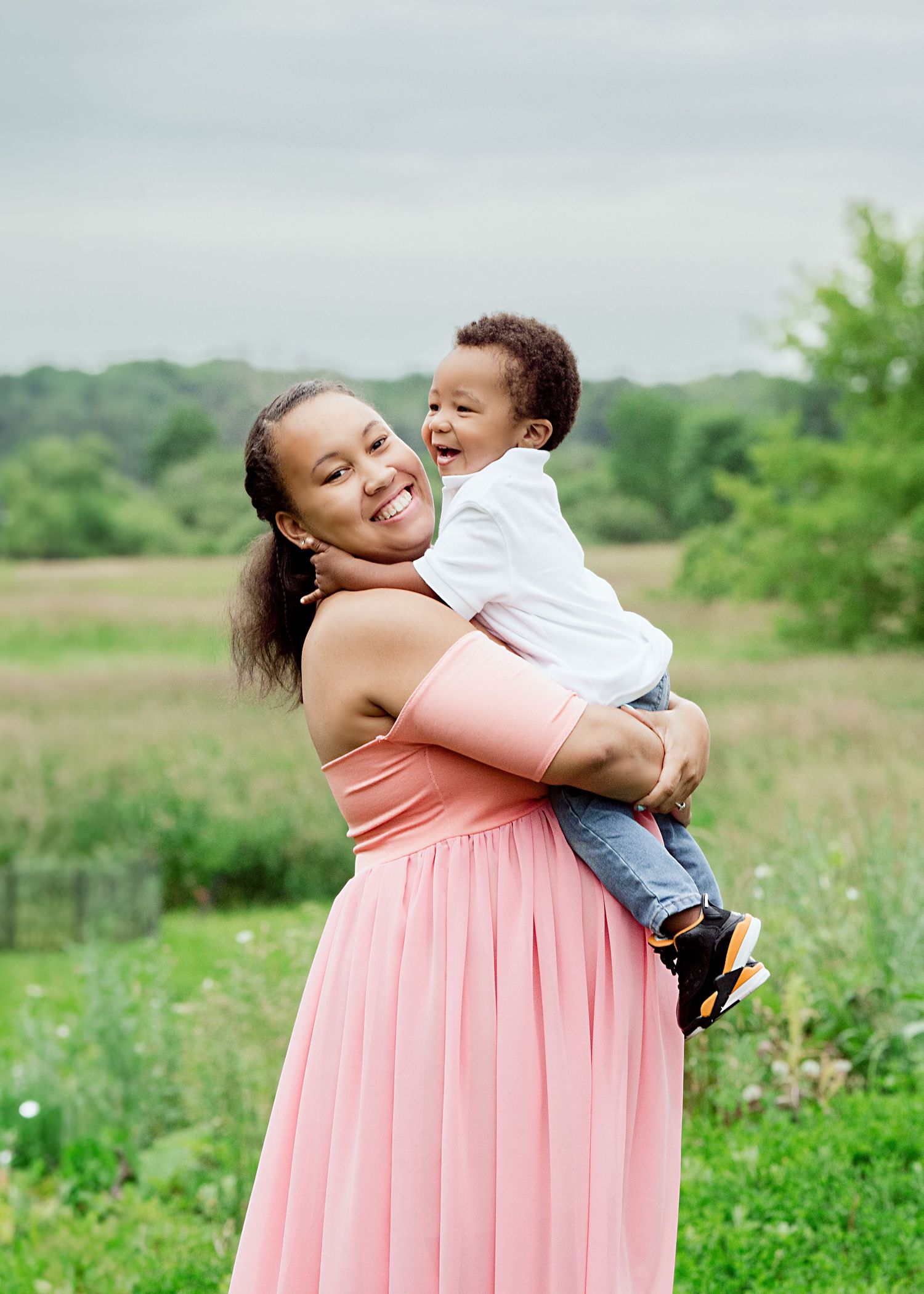Mother in coral dress joyfully holds and plays with toddler in outdoor setting.