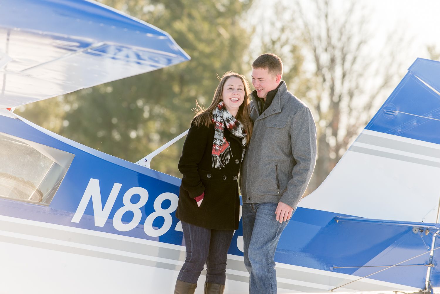 A couple poses together next to a blue and white private airplane on a bright day.