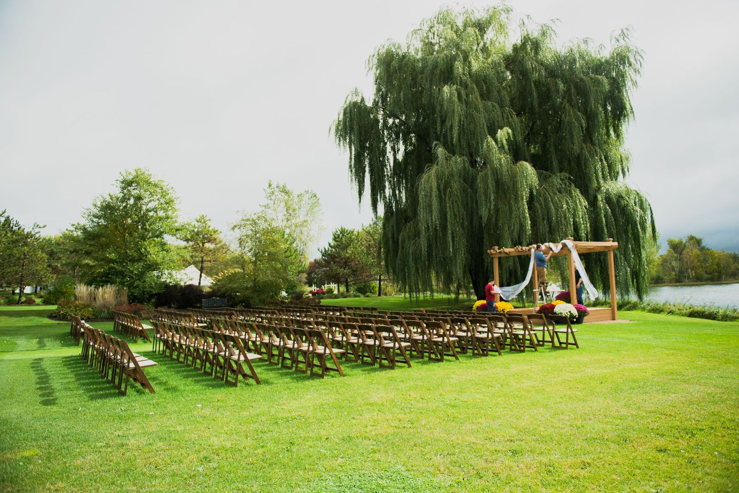 Outdoor wedding ceremony setup with wooden chairs on green lawn beneath a majestic weeping willow tree by the water. Andover wedding at Mage