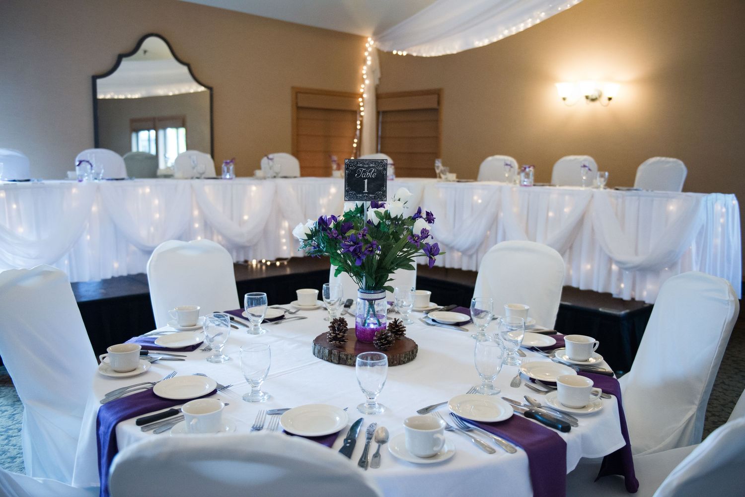 Formal dining setup with white chair covers, purple accents, and floral centerpiece in elegant banquet room. Andover wedding at Magestic Oak
