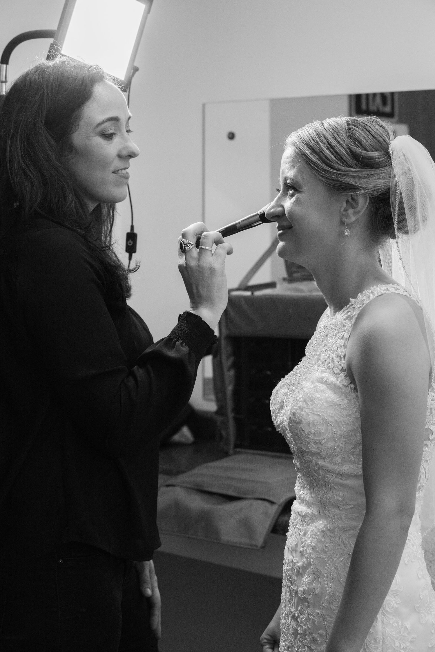 A makeup artist applies finishing touches to a bride's makeup in an intimate black and white pre-wedding preparation moment. Andover wedding