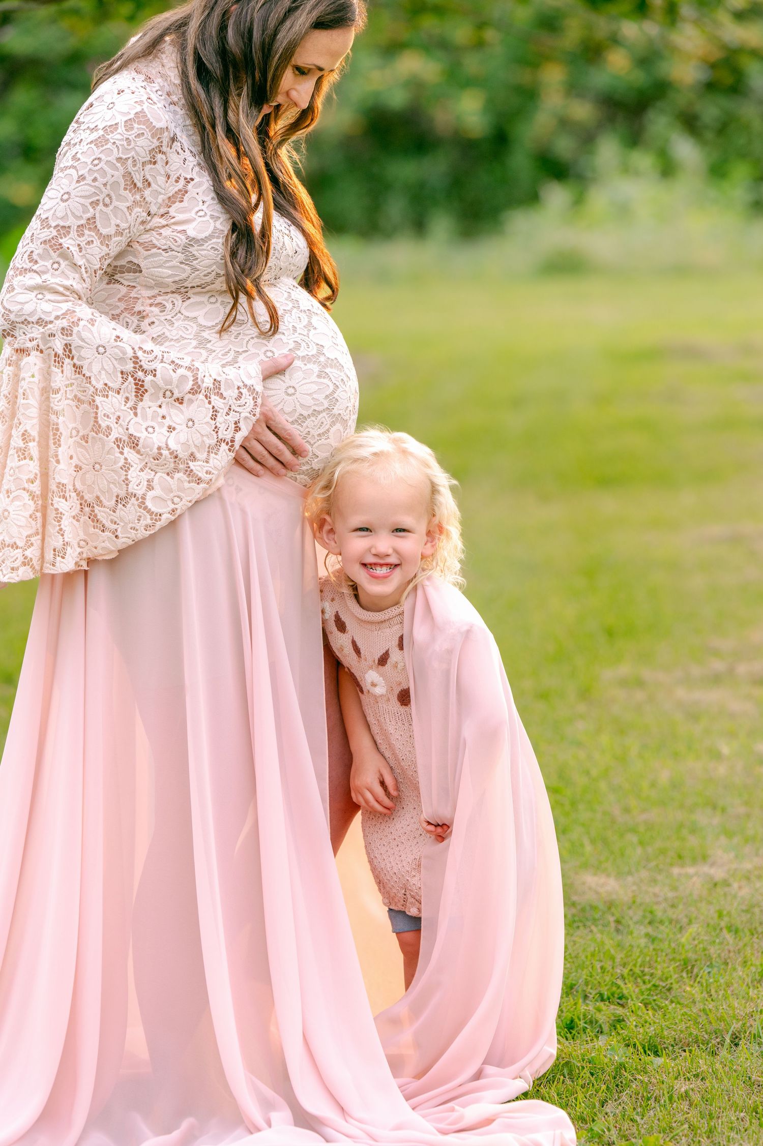 Mother in pink dress shares tender moment with child during outdoor maternity session.