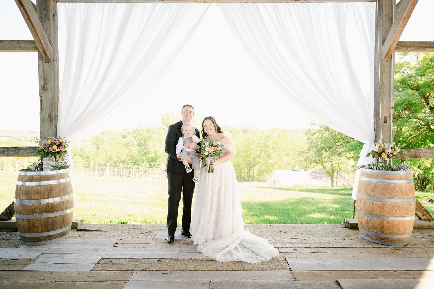 Wedding couple stands under draped pergola with rustic wine barrels and countryside backdrop.