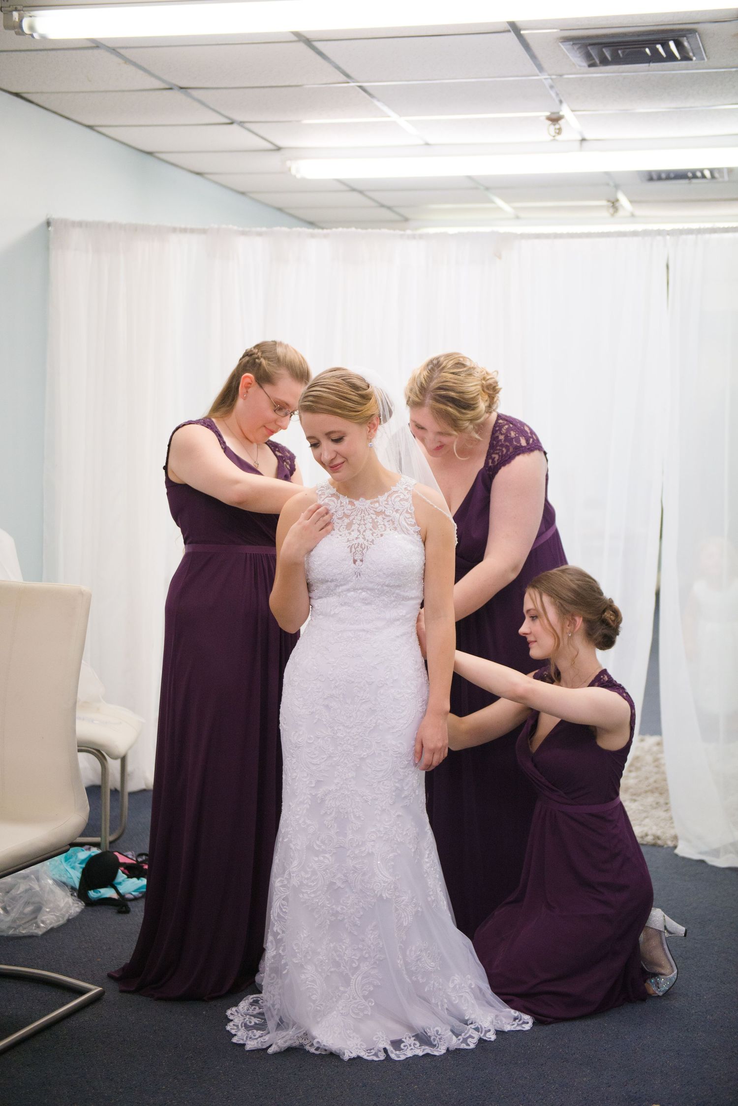 Bridesmaids in purple dresses help arrange a bride's lace wedding gown during pre-ceremony preparations. Andover wedding at Magestic Oaks Go