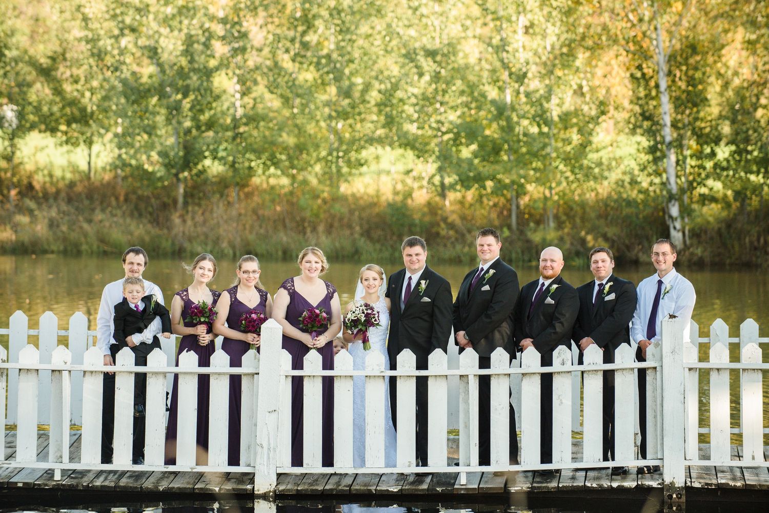 Wedding party poses together on a white picket fence bridge with autumn trees in background. Andover wedding at Magestic Oaks Golf Club