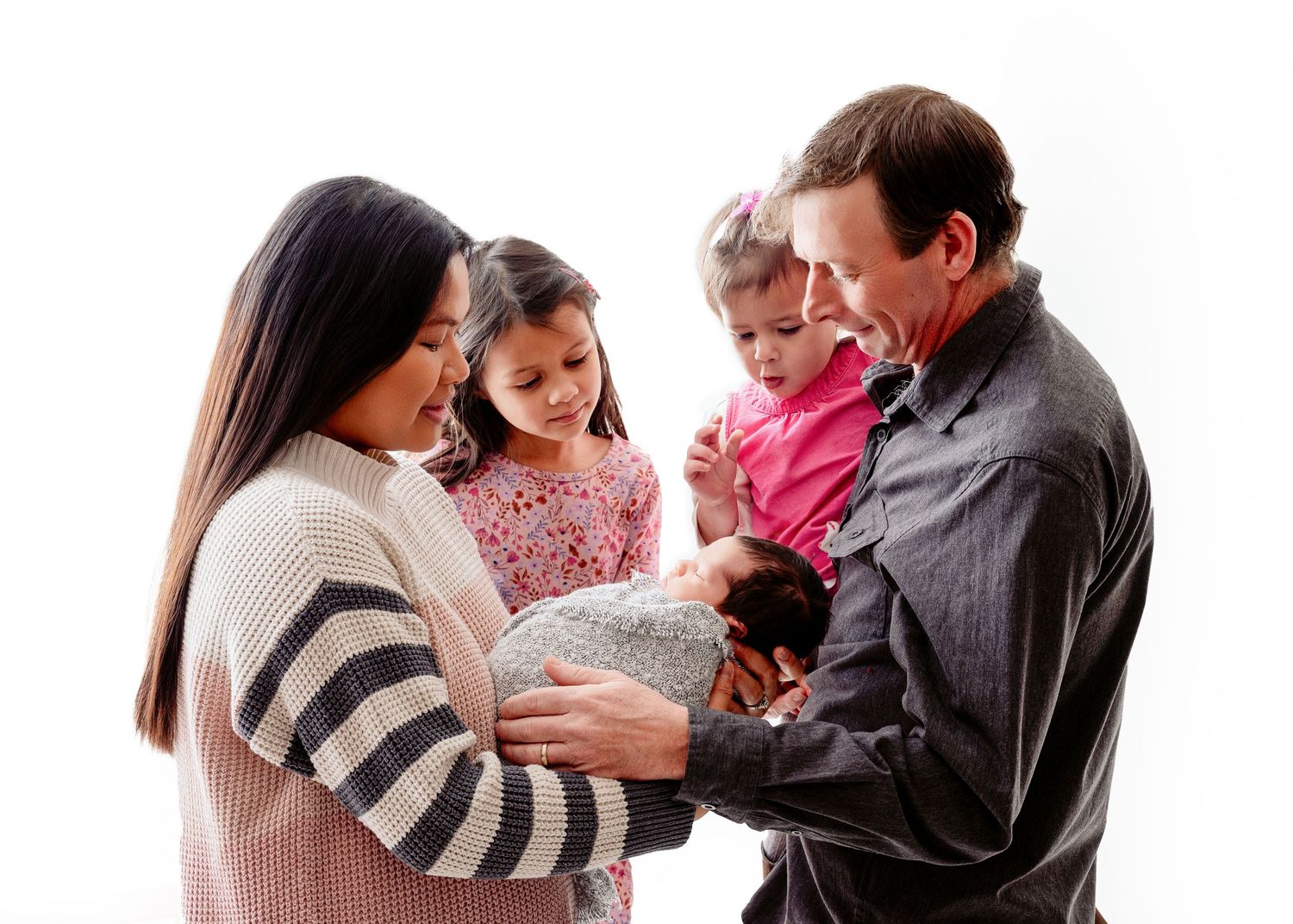 A group stands together looking lovingly at a small newborn baby wrapped in a blanket against a white background.
