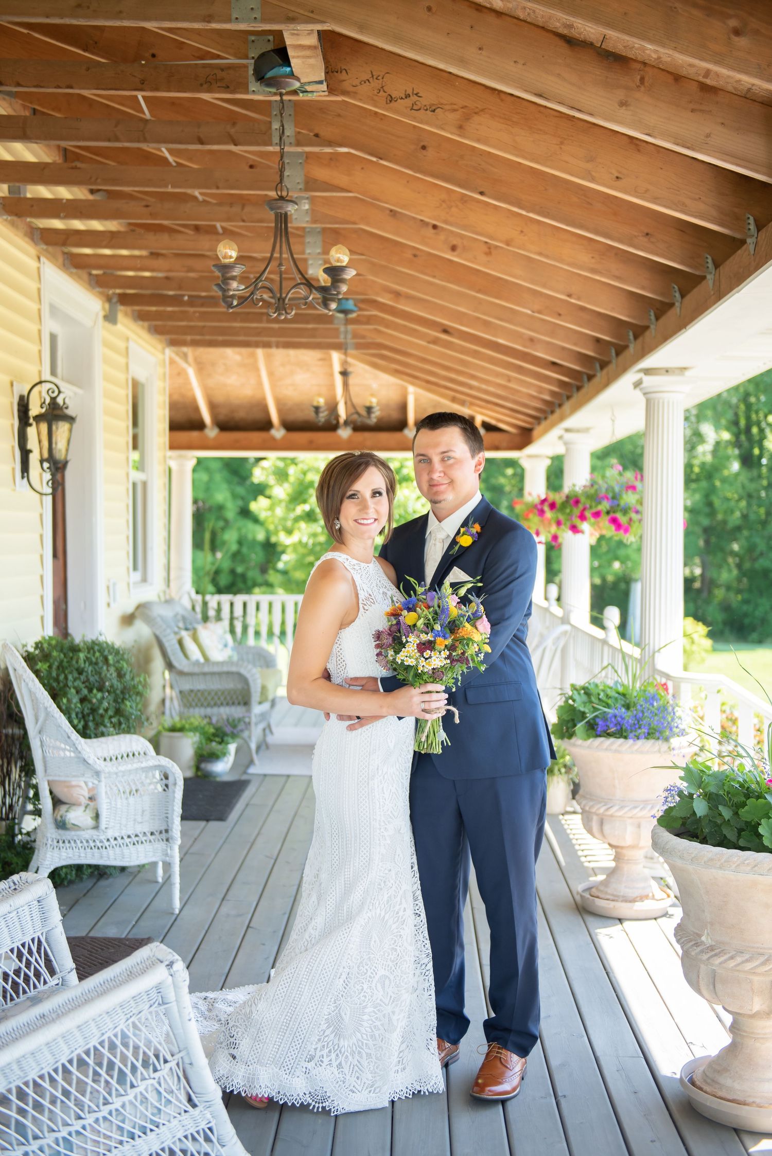 A couple stands on a covered porch with white wicker furniture and hanging flower baskets on their wedding day.