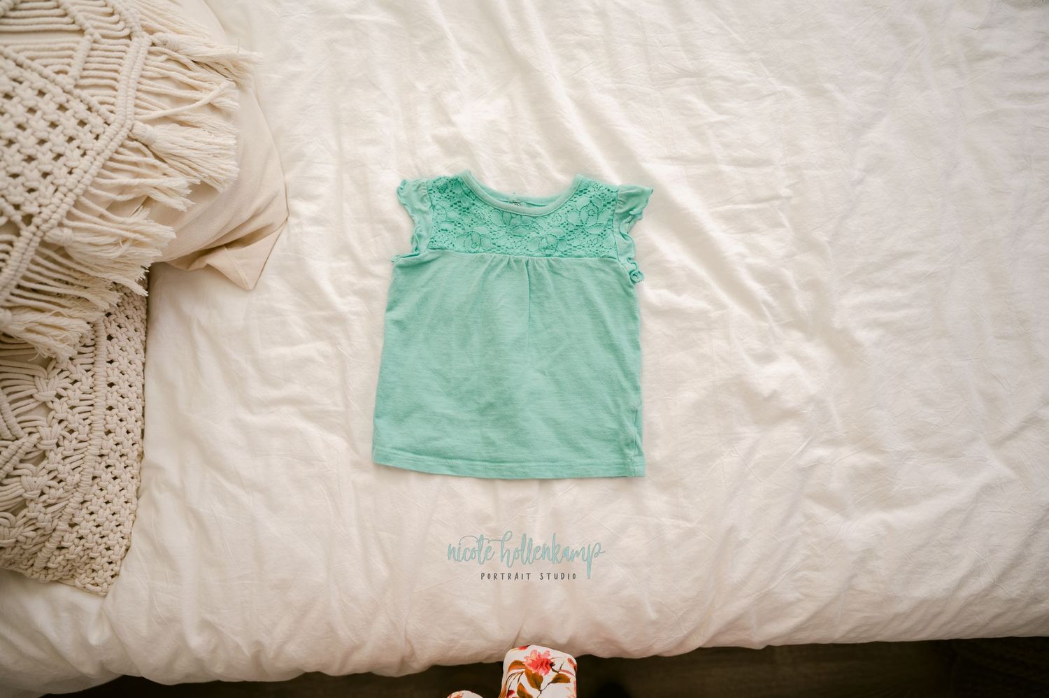 A mint green sleeveless baby shirt laid flat on a white bedspread next to a knit blanket.