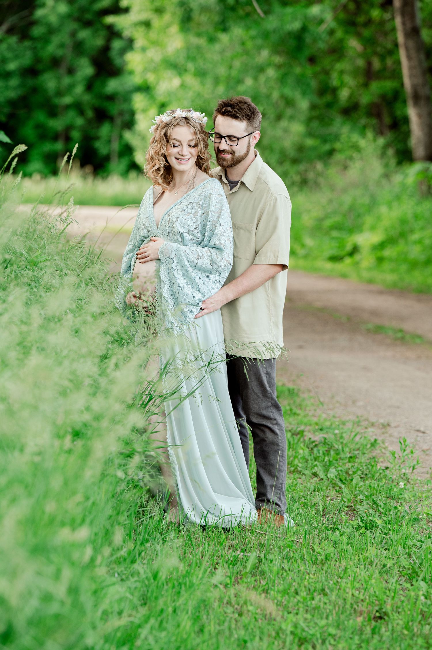 Expectant couple poses together on a nature path while wearing a flowing mint dress and casual attire.
