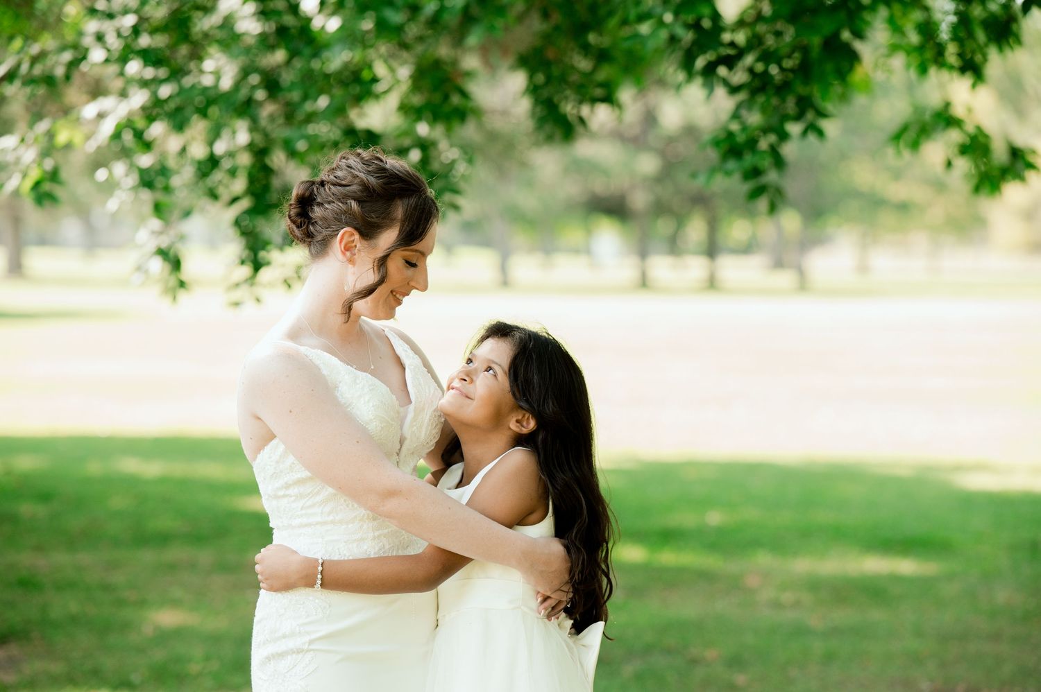A tender embrace between two figures in white dresses on a sunny lawn surrounded by green trees.