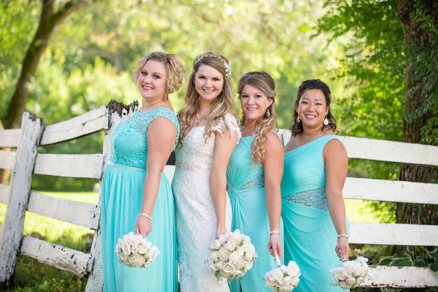 Bridal party holding bouquets poses by white fence in matching turquoise dresses. Rum River Barn Milaca Minnesota Weddings