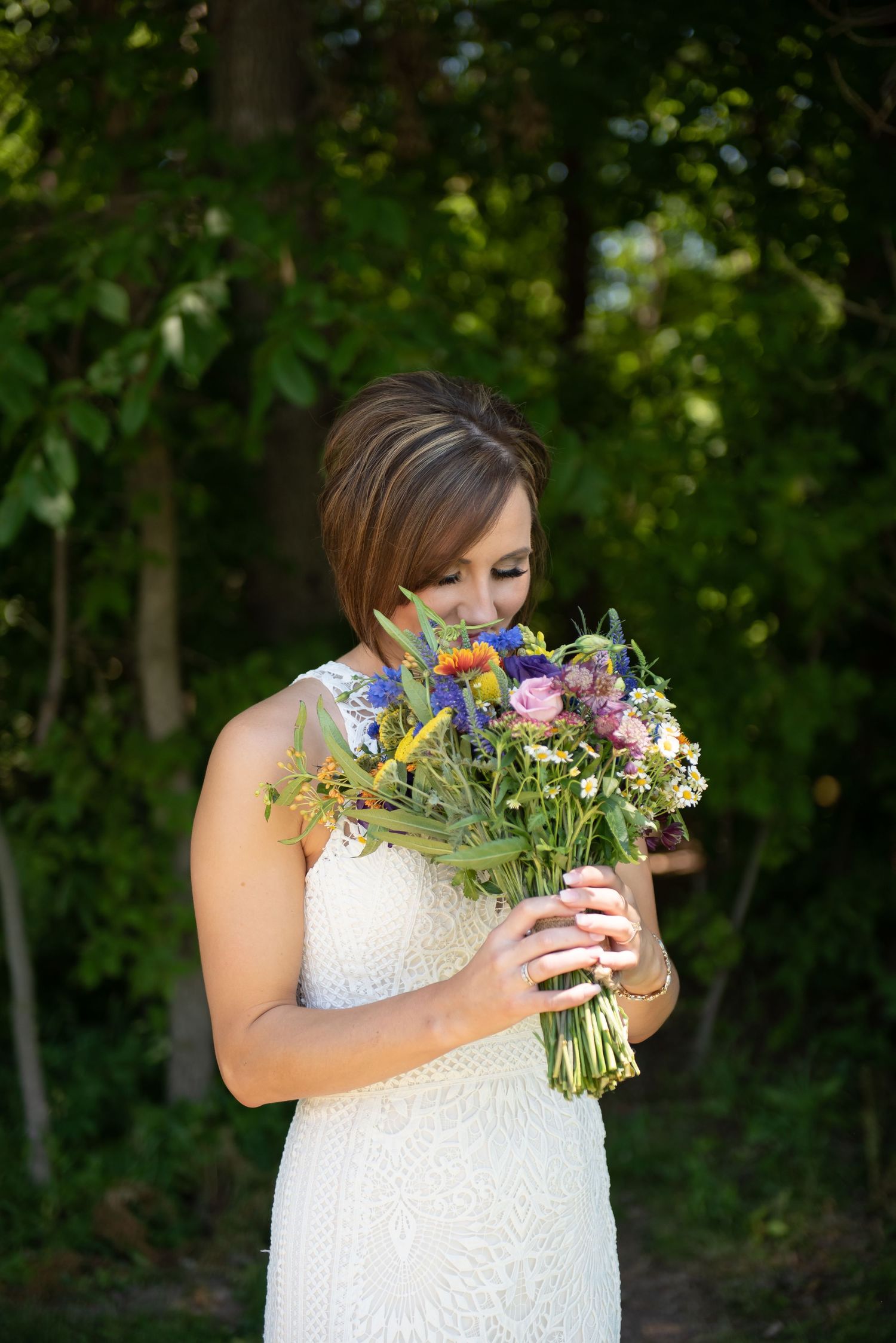 Person in white lace dress holds a vibrant bouquet of pink and blue flowers against green foliage background.