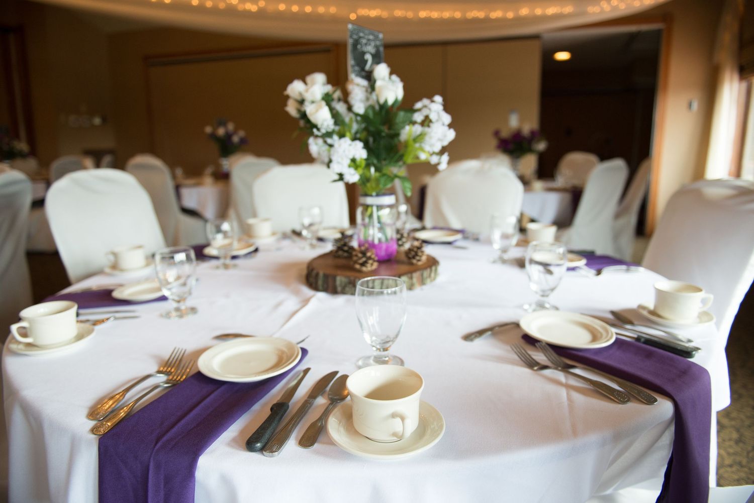 Elegant round banquet table with white linens, purple runner, and white floral centerpiece in formal dining room setting. Andover wedding at