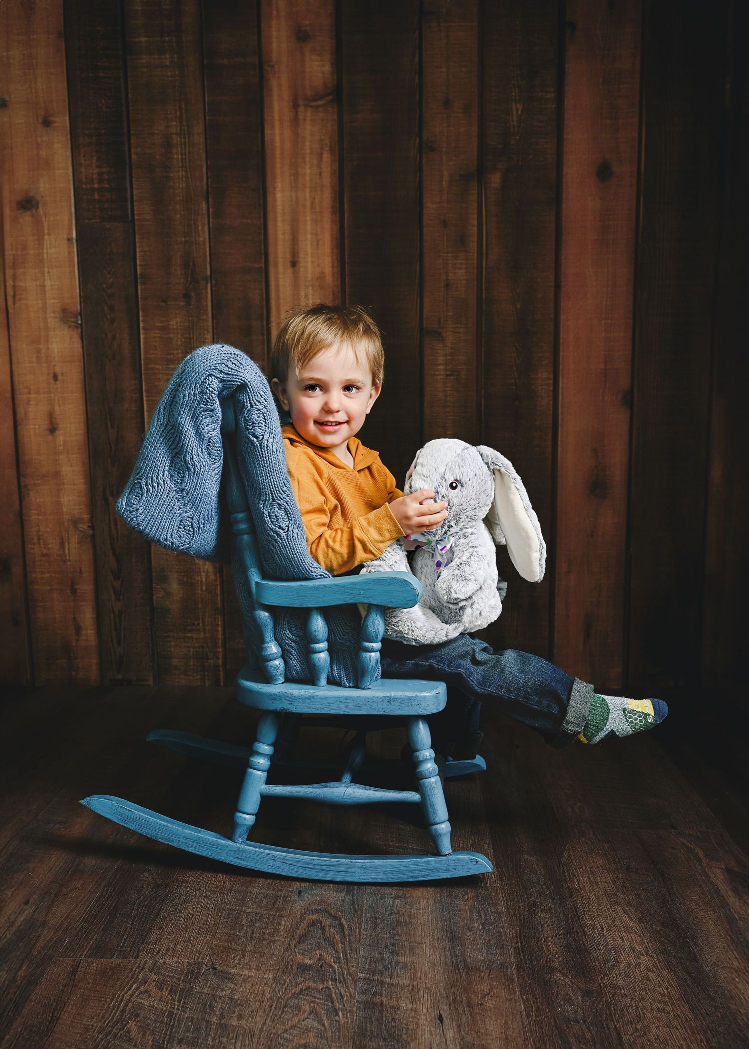 A child relaxes in a blue rocking chair while cuddling a soft gray bunny stuffed animal against wooden paneling.