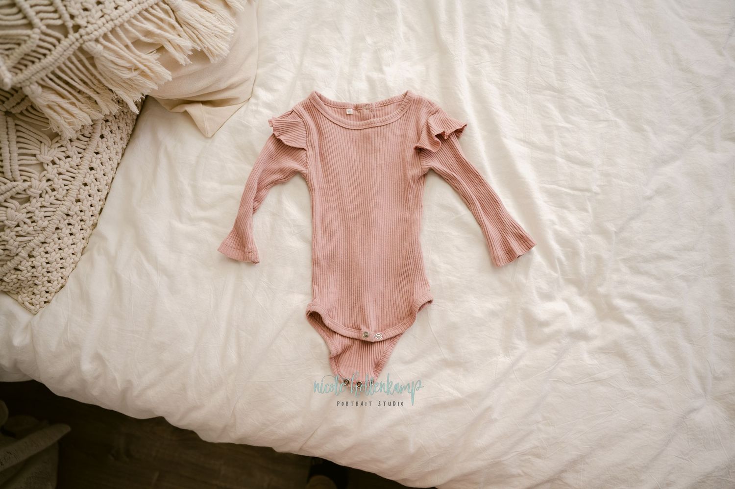 Pink long-sleeved baby onesie laid out on white bedding with knit blanket in corner.
