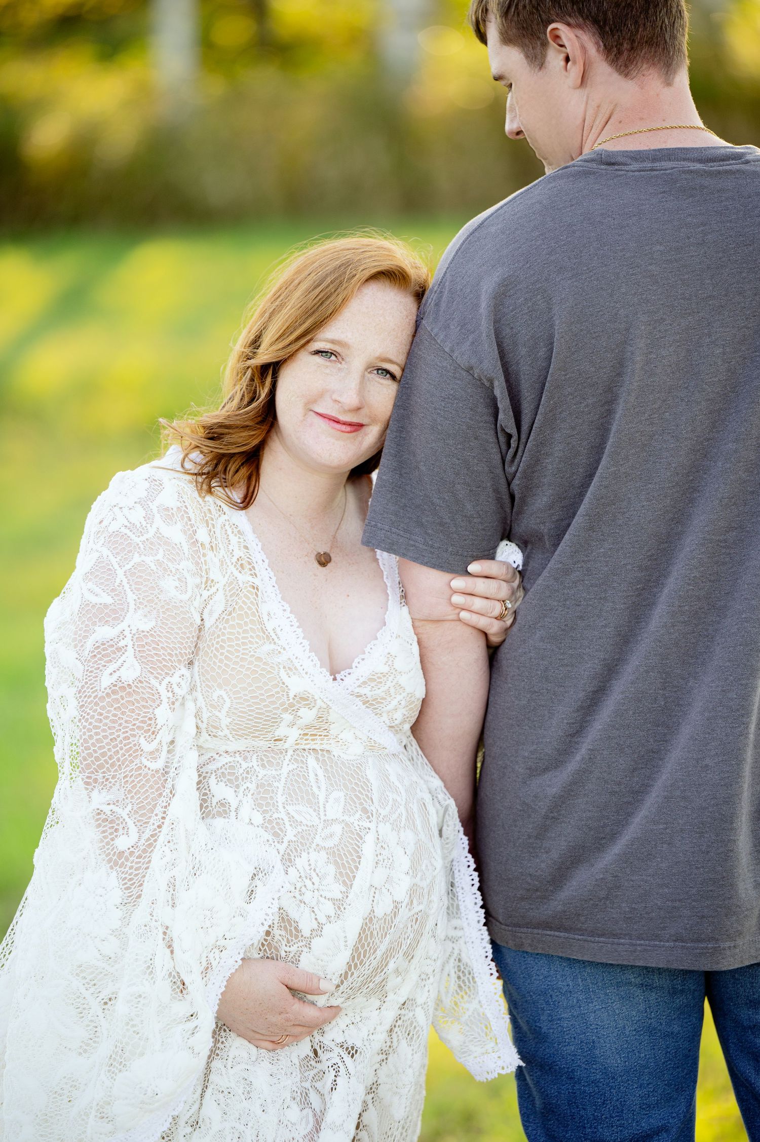A pregnant woman in a white lace dress holds her partner's arm while looking at the camera in an outdoor setting.