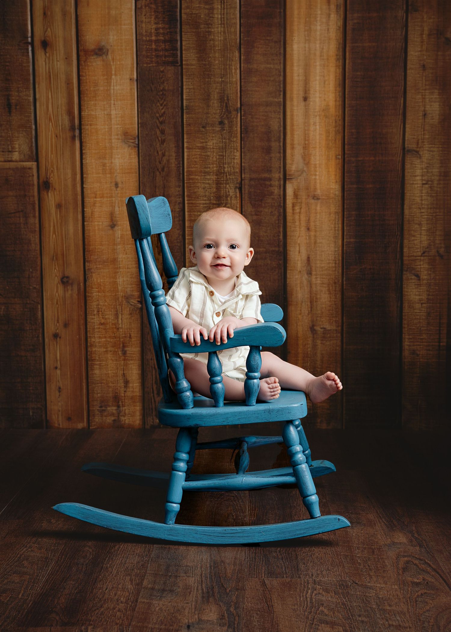 Baby in white outfit sitting in turquoise blue rocking chair against wooden background.