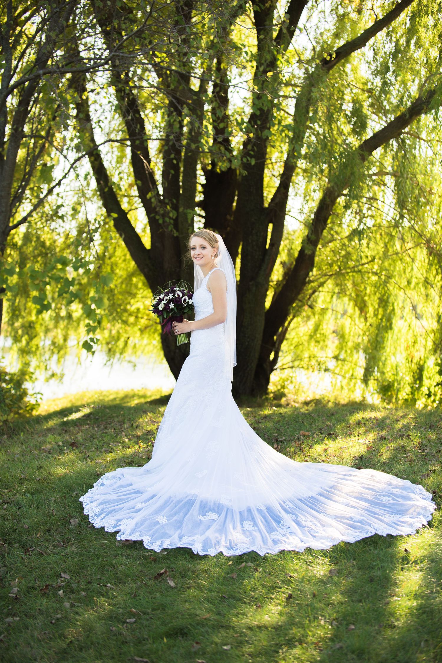 White wedding dress with a flowing train spreads beautifully across green grass under willow trees. Andover wedding at Magestic Oaks Golf Cl