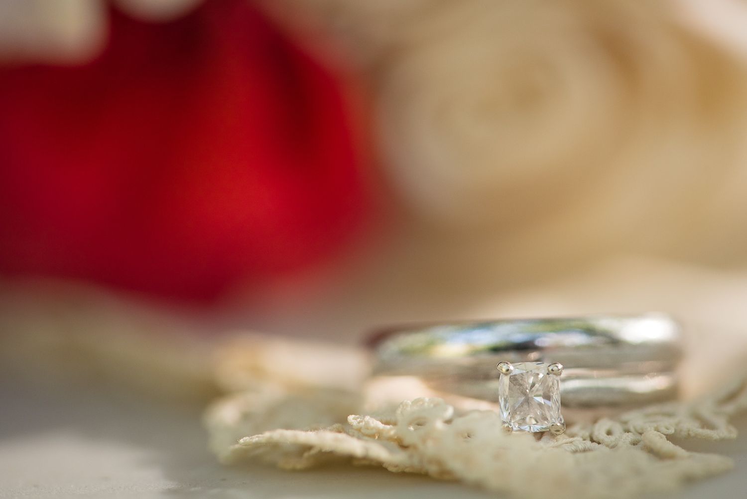 A delicate silver engagement ring with a small diamond rests on soft cream fabric with a blurred red background.