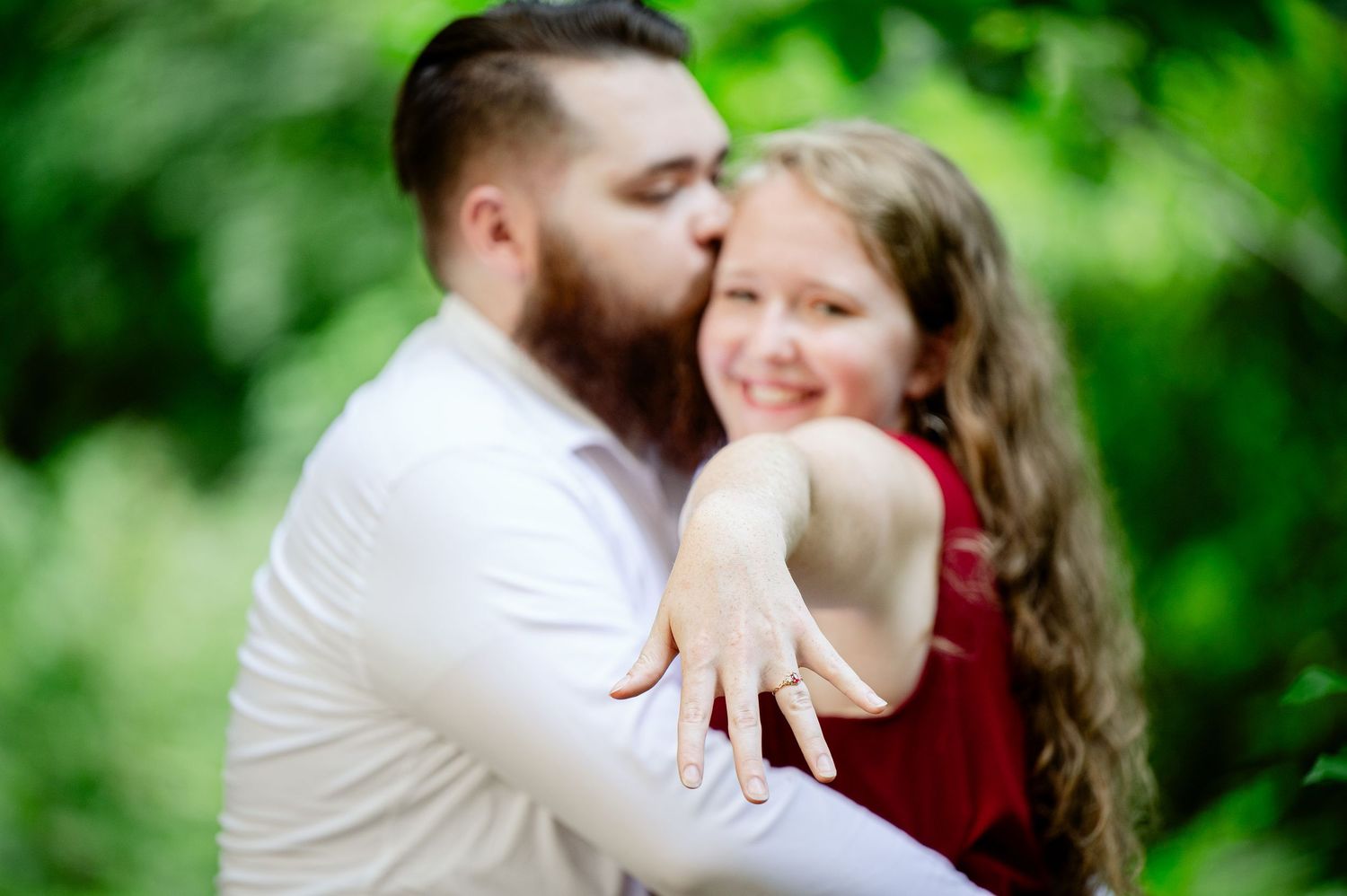 A couple embraces outdoors in a romantic engagement photo with a blurred green natural background.
