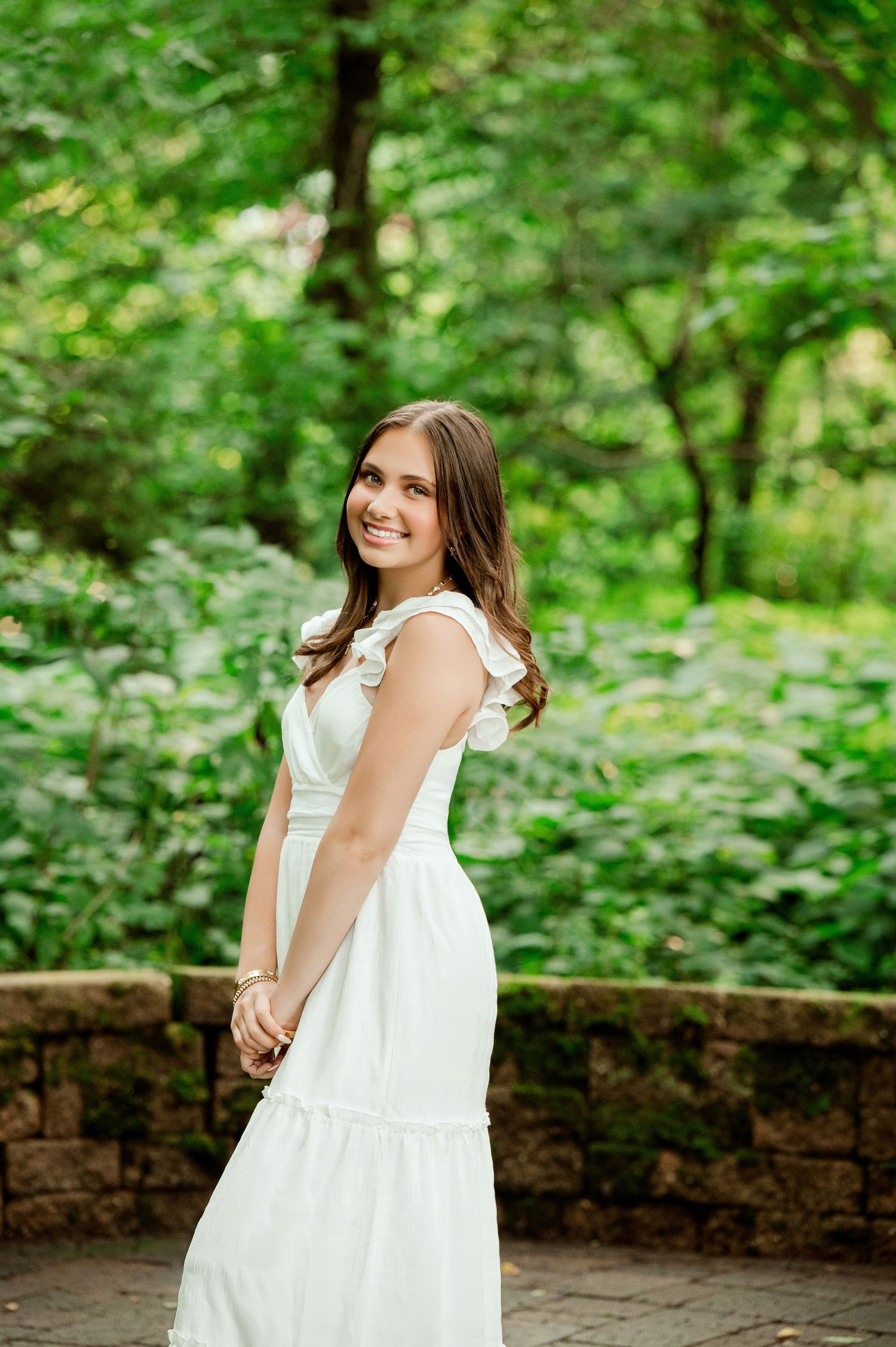 Person in white dress poses against stone wall with lush green foliage in background.