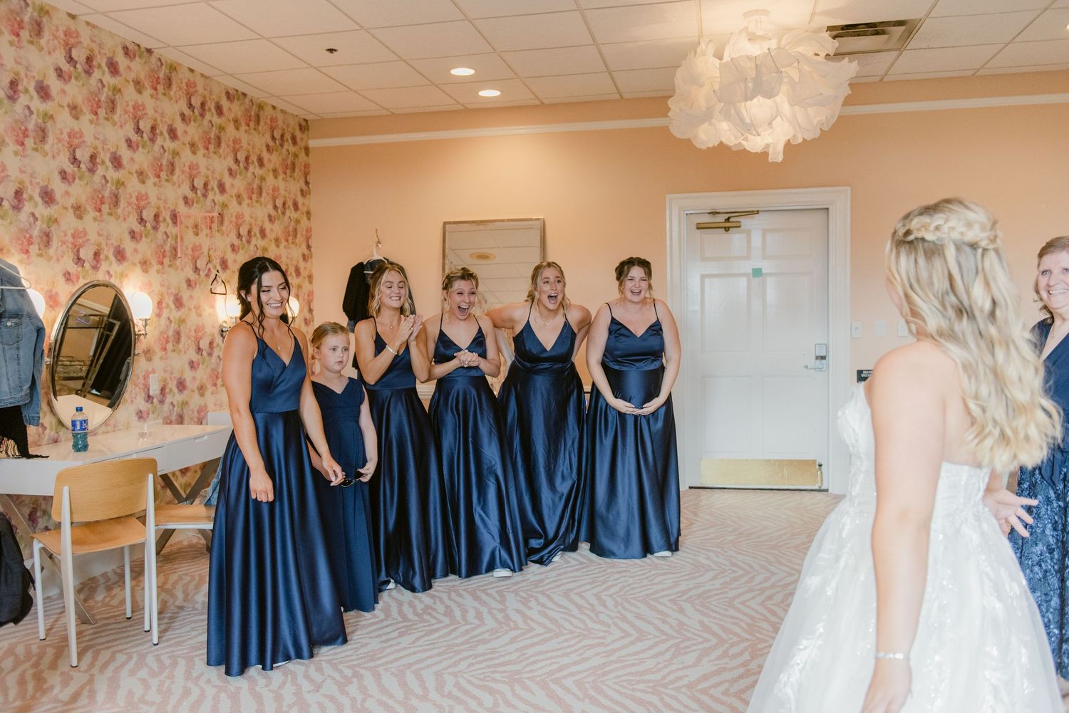 Group wearing matching navy blue satin bridesmaid dresses stand in an elegant dressing room with floral wallpaper.