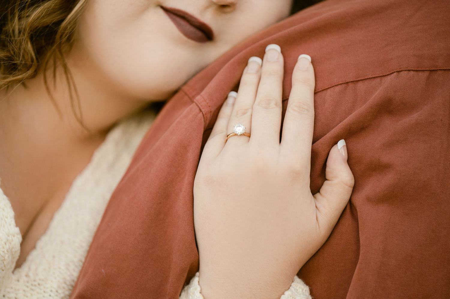Close-up of a hand wearing a diamond engagement ring resting on rust-colored fabric against cream lace. St cloud area engagement photos.