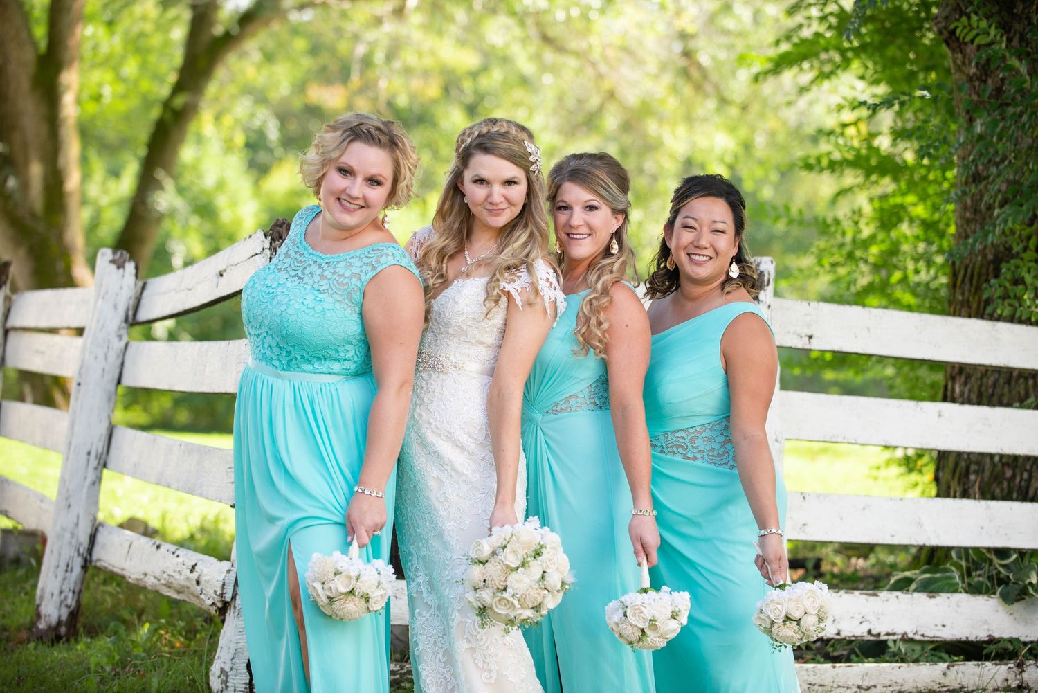 A bridal party wearing turquoise bridesmaid dresses and white wedding gown pose together by a white fence outdoors. Rum River Barn Milaca Mi