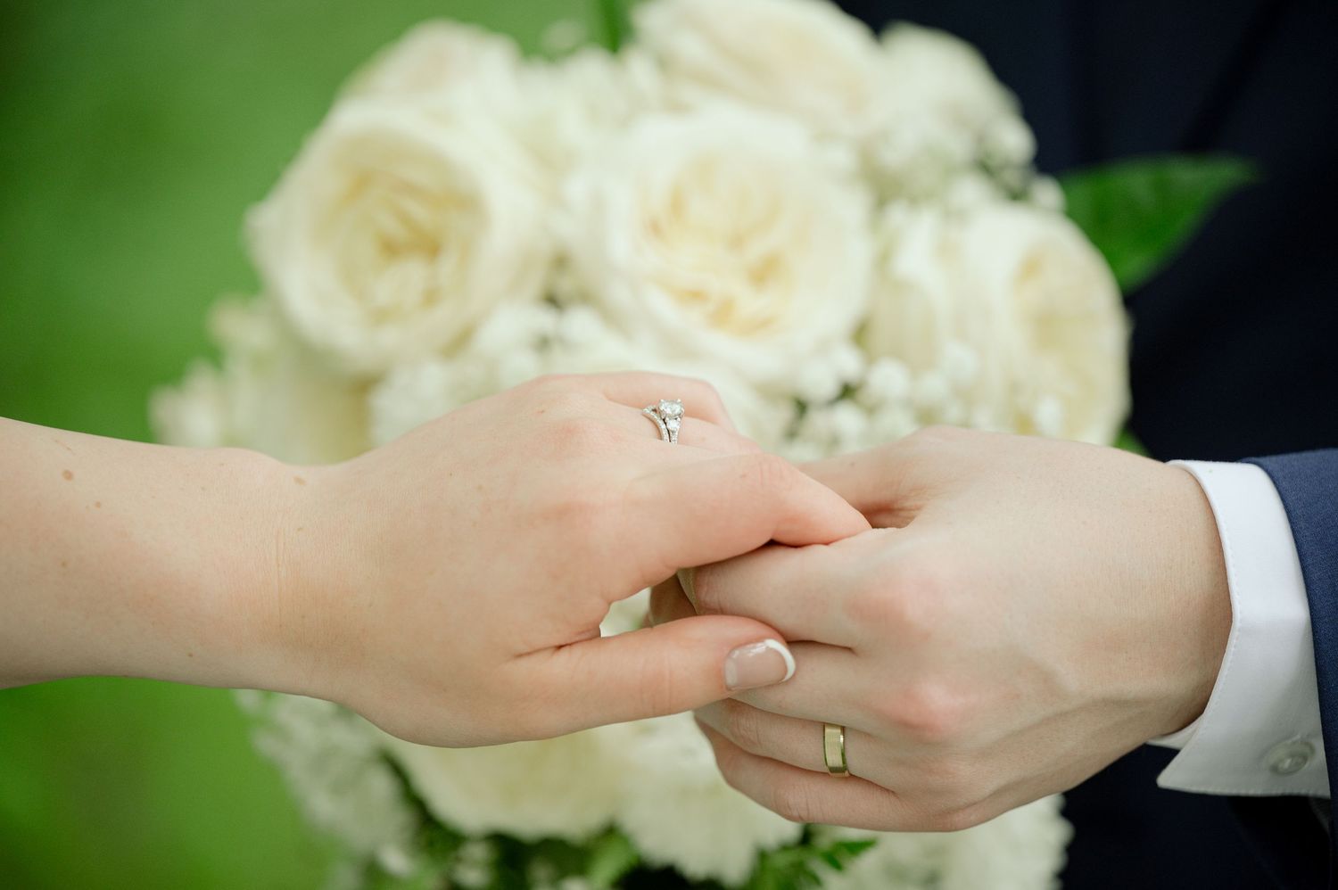 Close up of two newlyweds' hands with wedding rings and white roses bouquet in the background.
