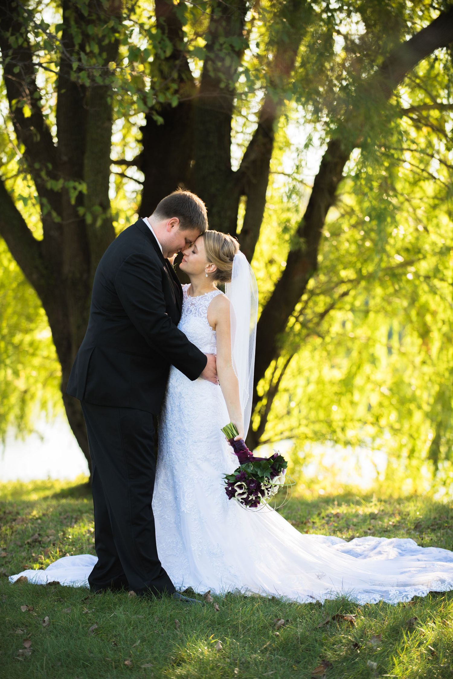 Intimate wedding moment captured under a majestic tree with flowing white dress trailing behind. Andover wedding at Magestic Oaks Golf Club