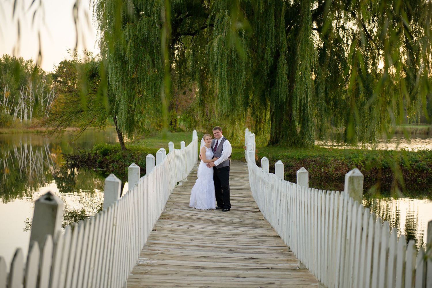 Couple standing on white wooden bridge with weeping willow trees and water in background. Andover wedding at Magestic Oaks Golf Club