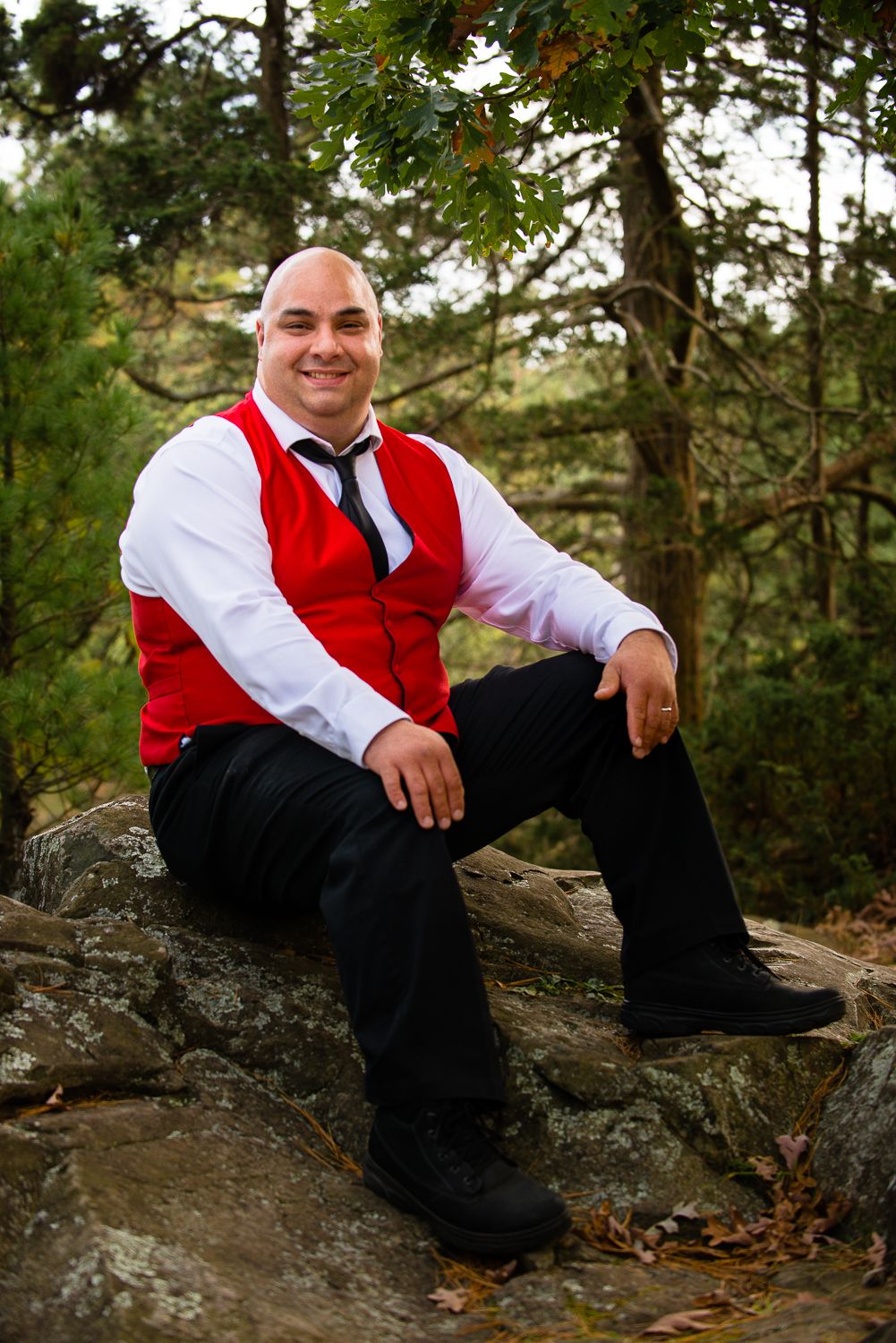 Person in formal attire wearing red vest and white shirt relaxes on rocky outcrop in outdoor forest setting.