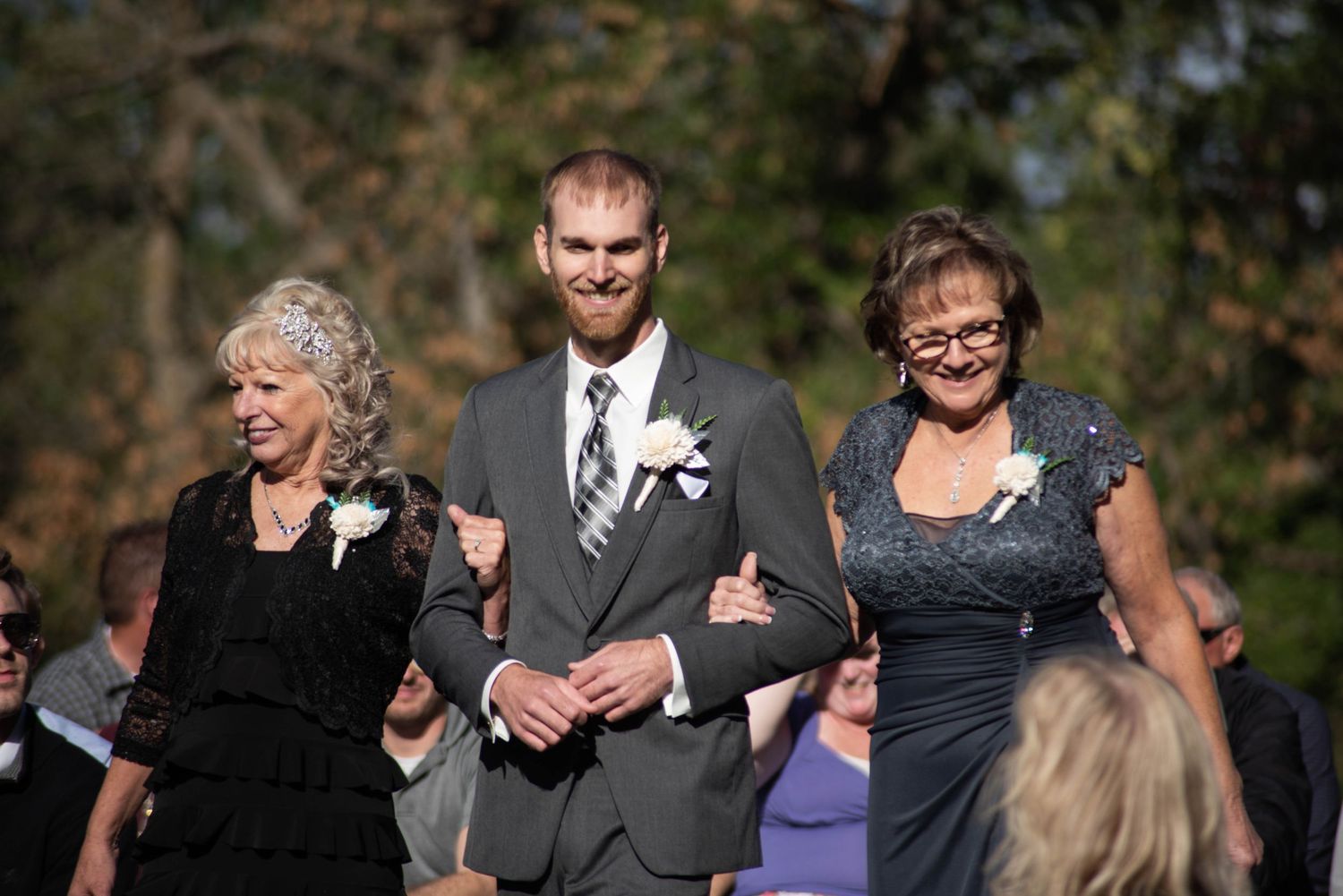 Three people walk arm in arm at an outdoor wedding ceremony on a sunny autumn day.
