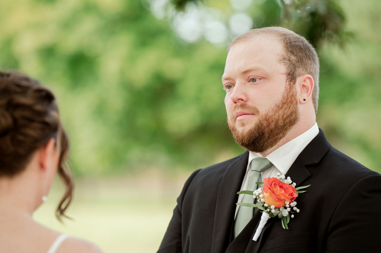 The Grands At Mulligans. Private vows Groom wearing black suit with coral boutonniere listens intently during outdoor wedding ceremony.