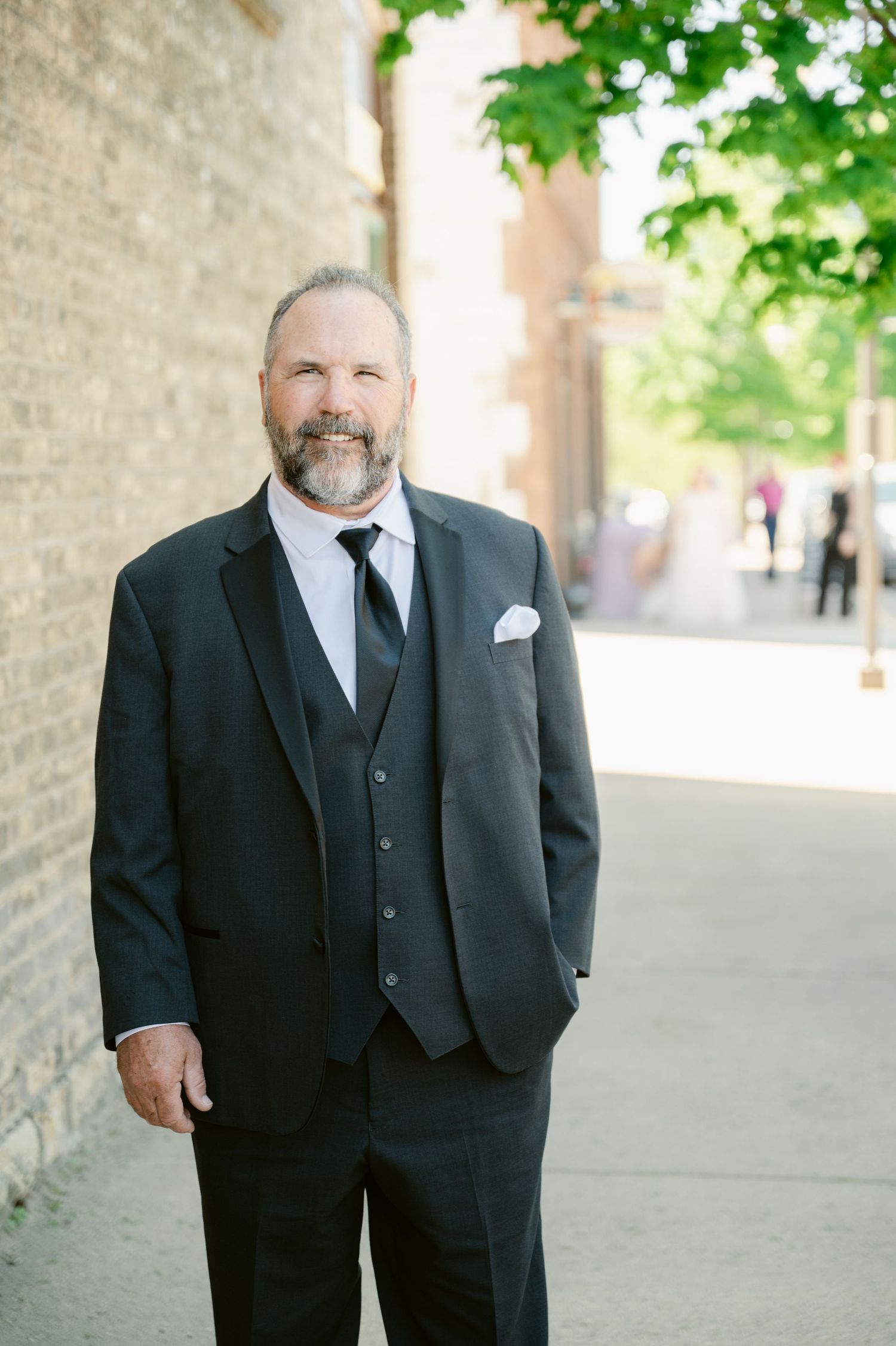 Formal portrait of wedding party member in classic black tuxedo standing against brick wall outdoors.