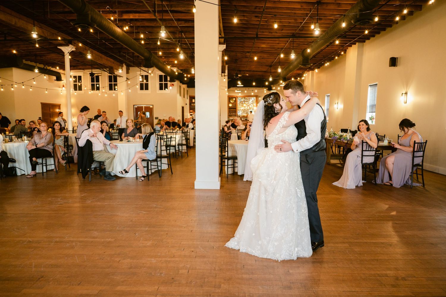 First dance moment in a rustic reception venue with string lights and wooden floors.
