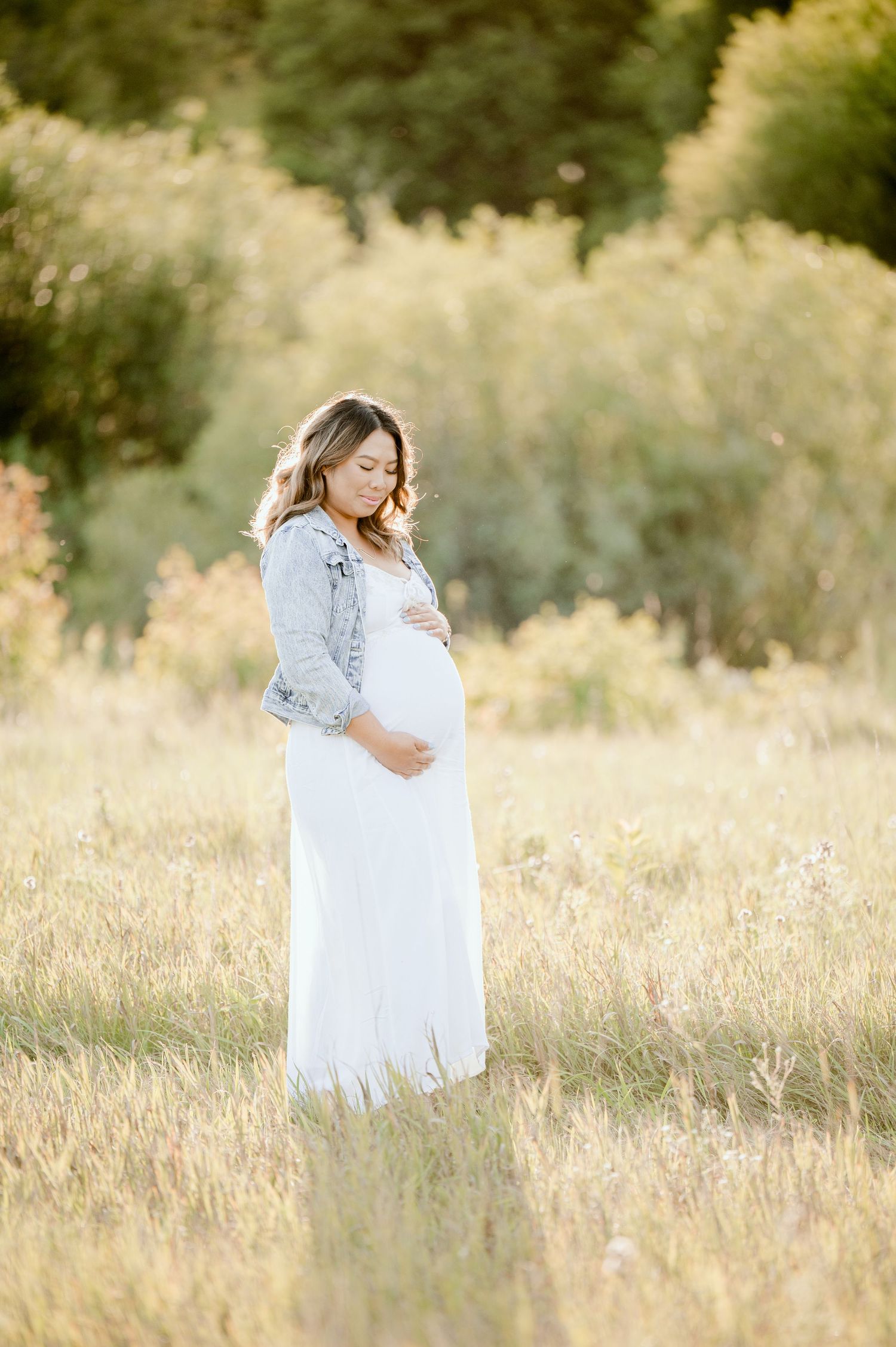 A pregnant woman in a flowing white dress and denim jacket stands in a sunlit golden field at sunset.