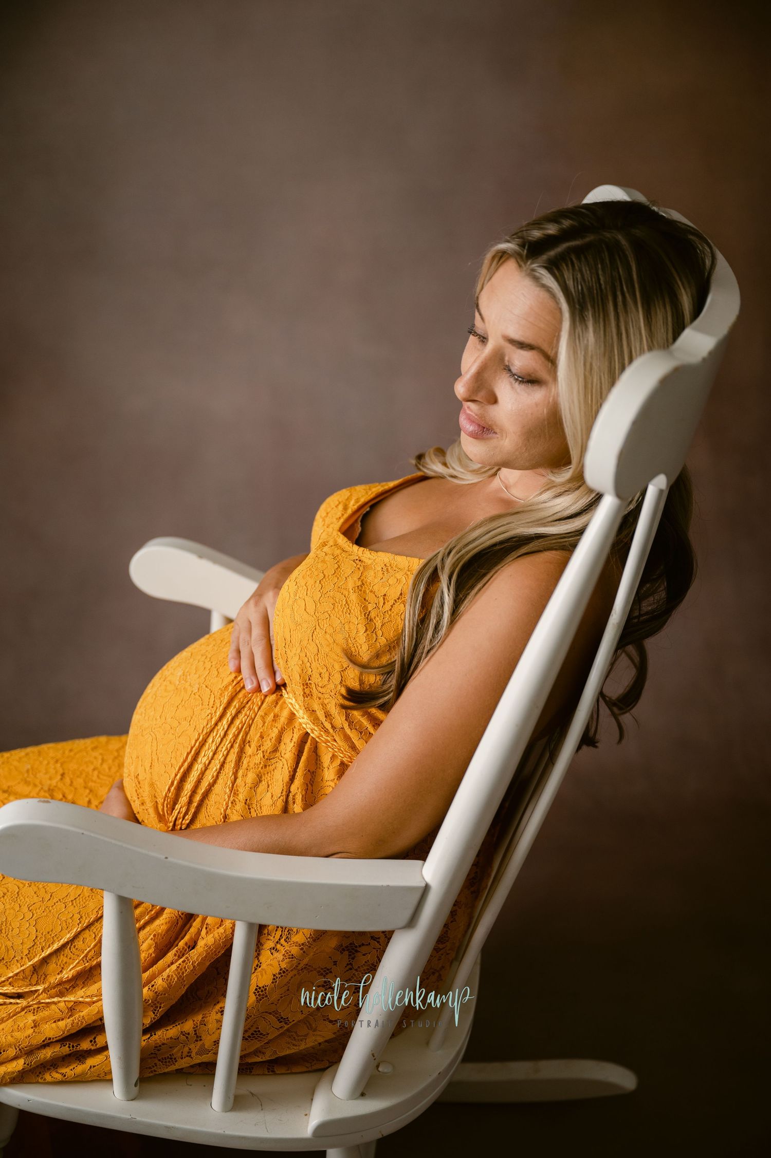 A pregnant woman in a yellow maxi dress relaxes peacefully in a white rocking chair against a brown backdrop.