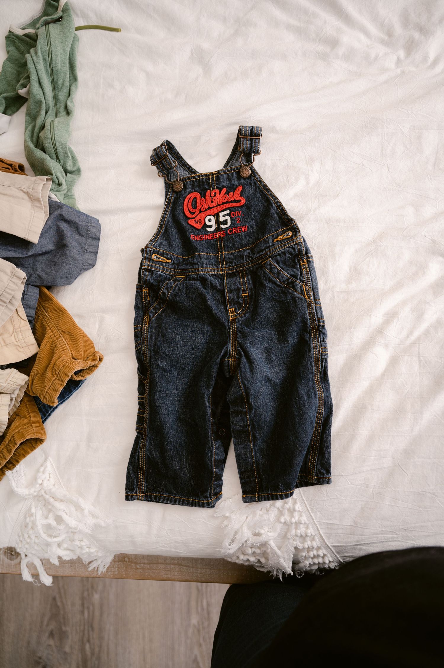 Small denim overalls with red '95' patch laid on white bedding next to scattered baby clothes.