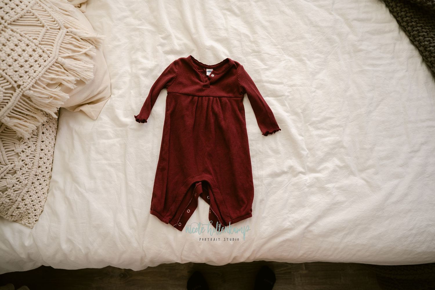 A burgundy red baby romper with long sleeves laid flat on a white bedding with a knit blanket in view.