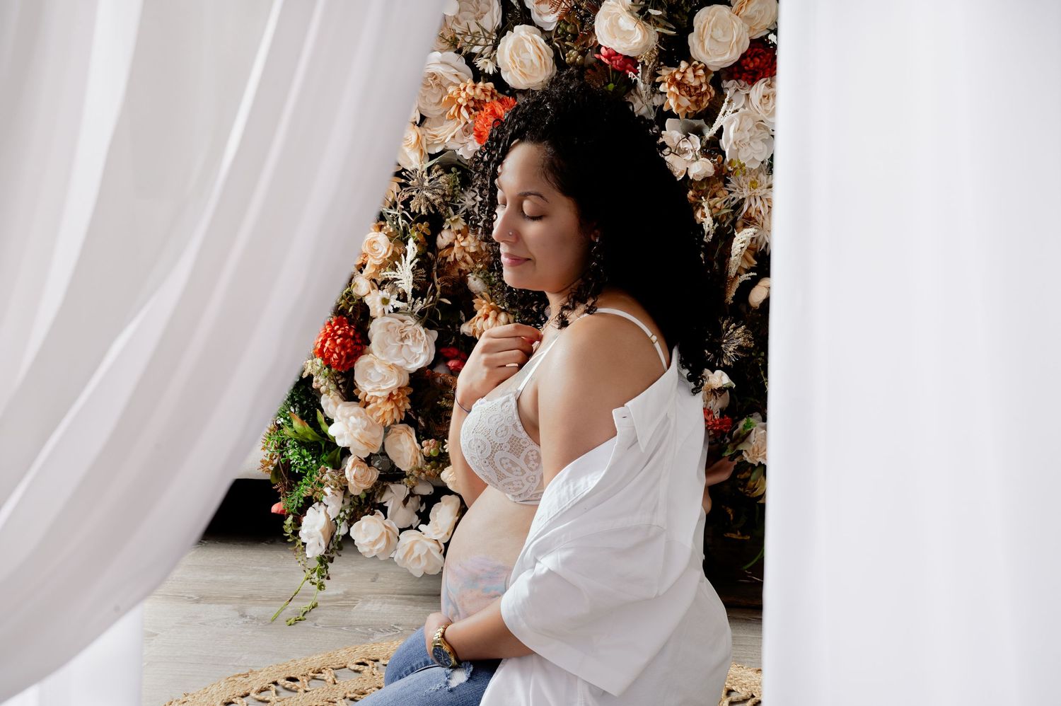 Expectant mother in white dress sitting beside floral wall installation.