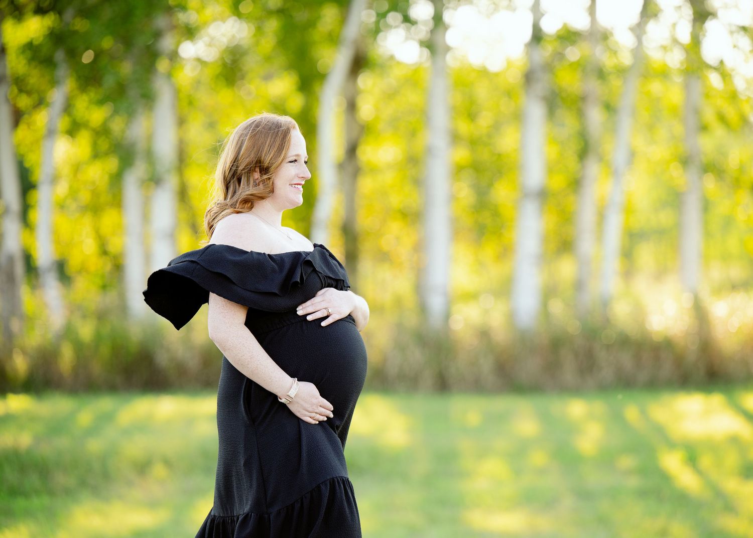 An expectant mother in an elegant black off-shoulder dress poses in front of sunlit aspen trees.