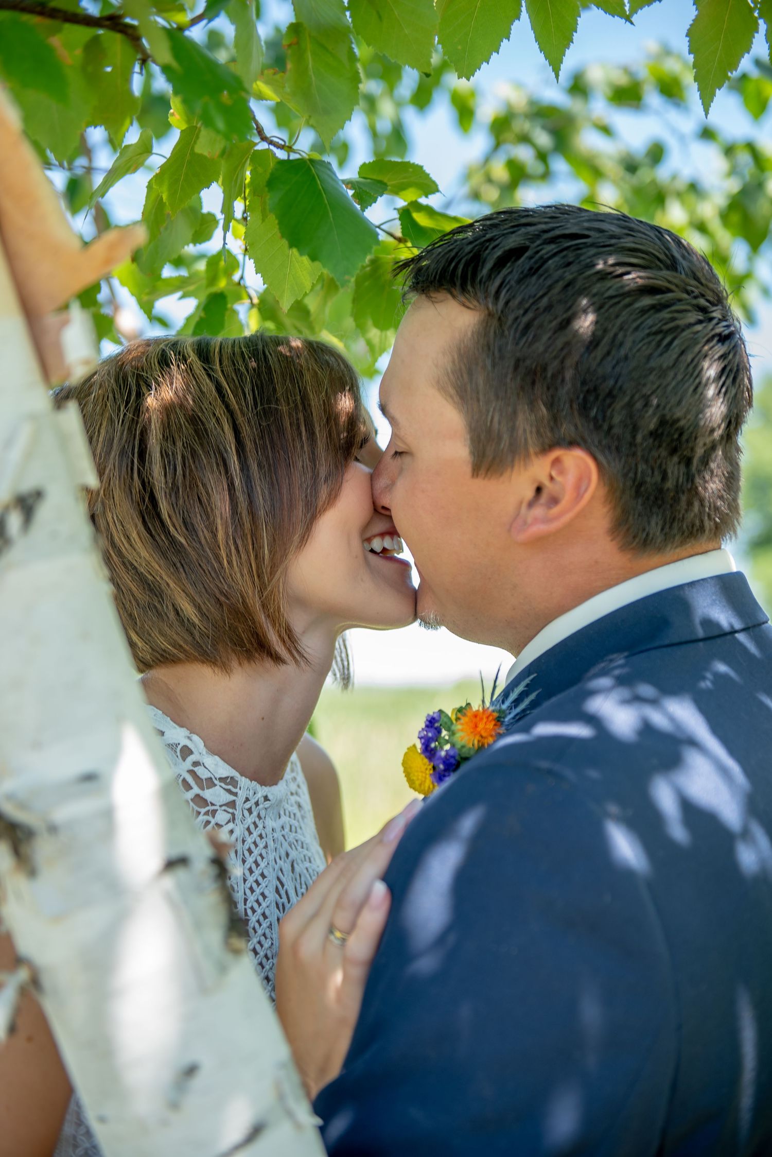 A tender wedding day kiss captured under green tree leaves with dappled sunlight.
