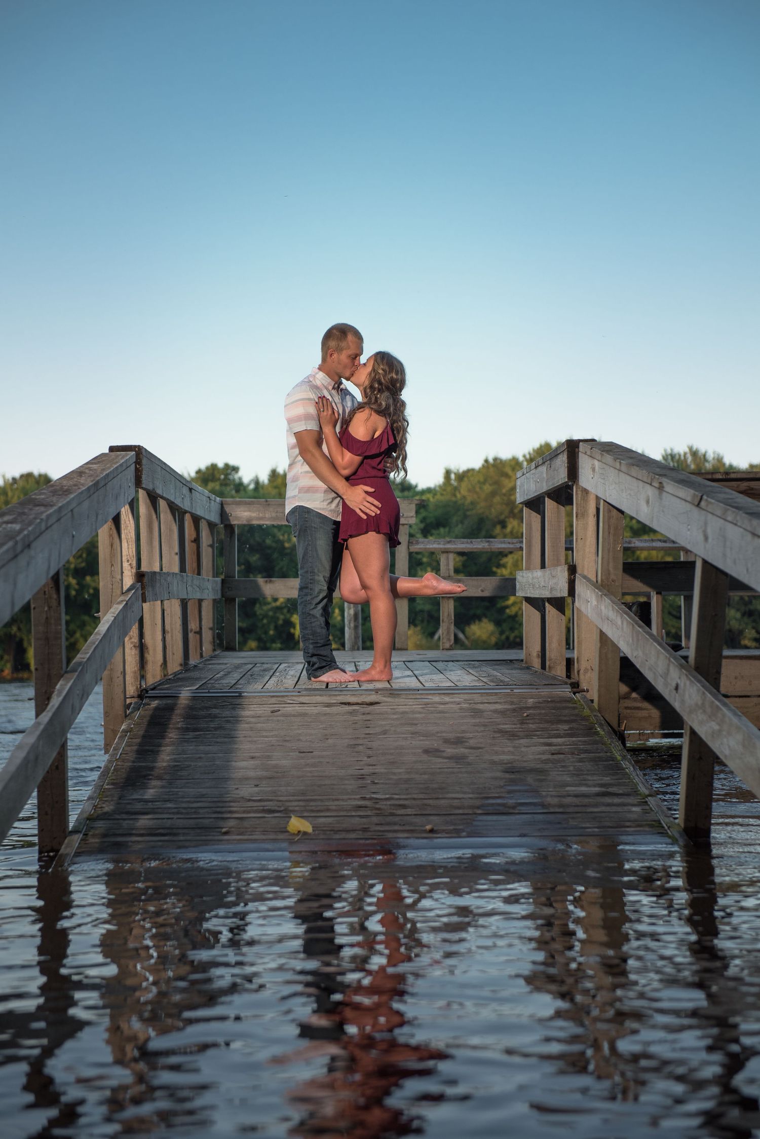 A romantic couple embraces on a wooden bridge over water during sunset, creating reflections below.
