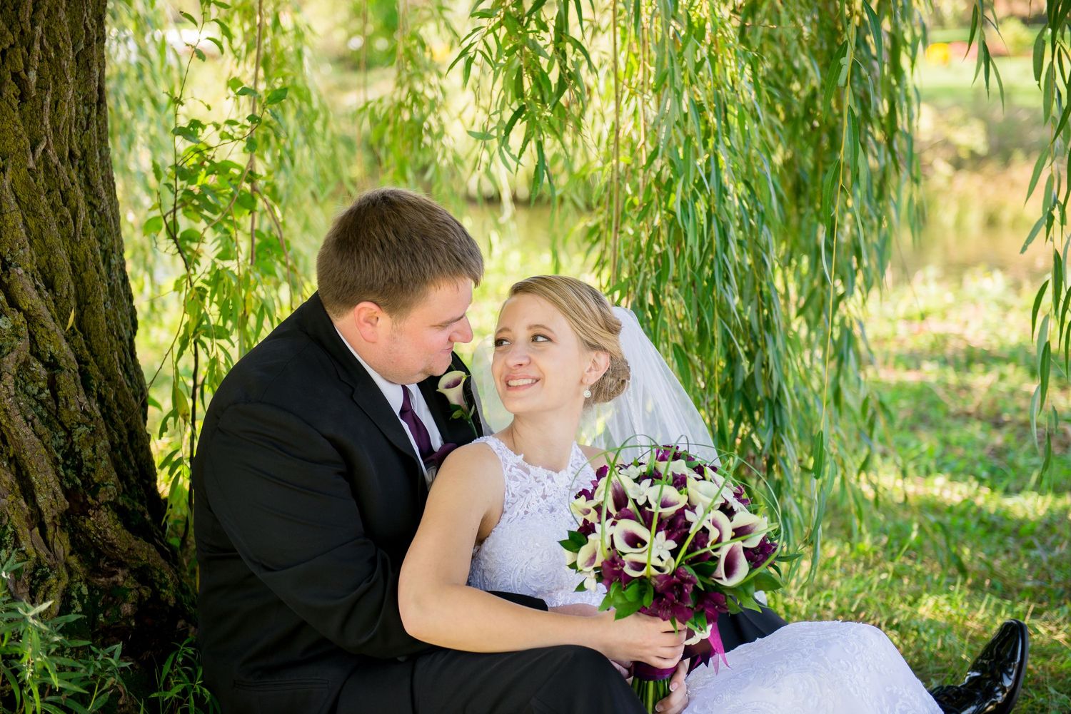 A romantic wedding photo under a weeping willow tree features a couple with a purple and white bouquet. Andover wedding at Magestic Oaks Gol
