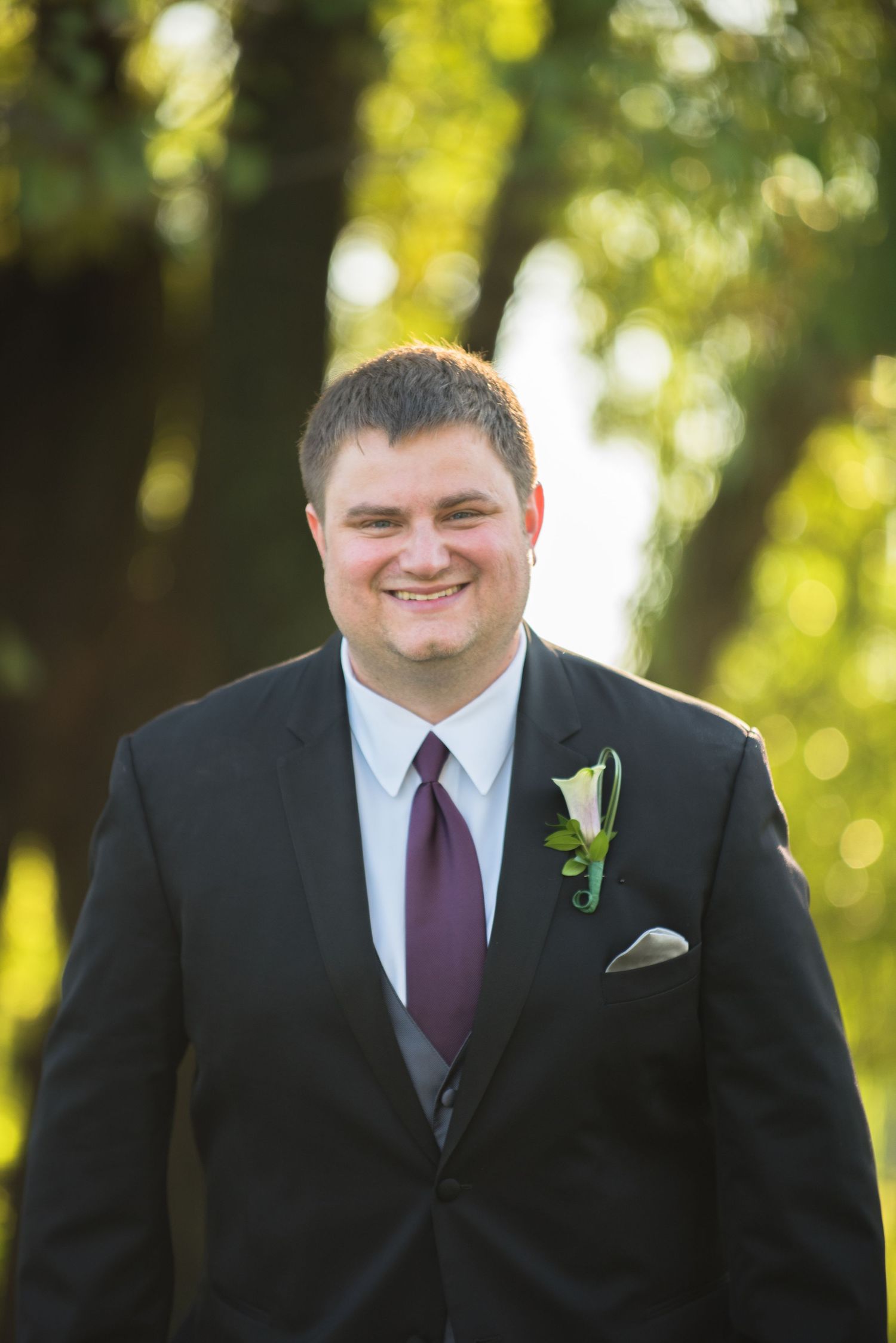 Groom in black tuxedo with purple tie and white boutonniere smiles outdoors. Andover wedding at Magestic Oaks Golf Club