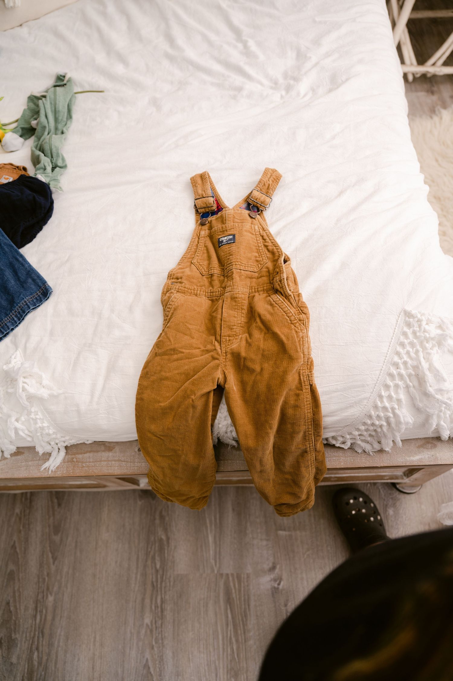 Brown toddler OshKosh overalls laid flat on white bedding against gray wooden flooring.