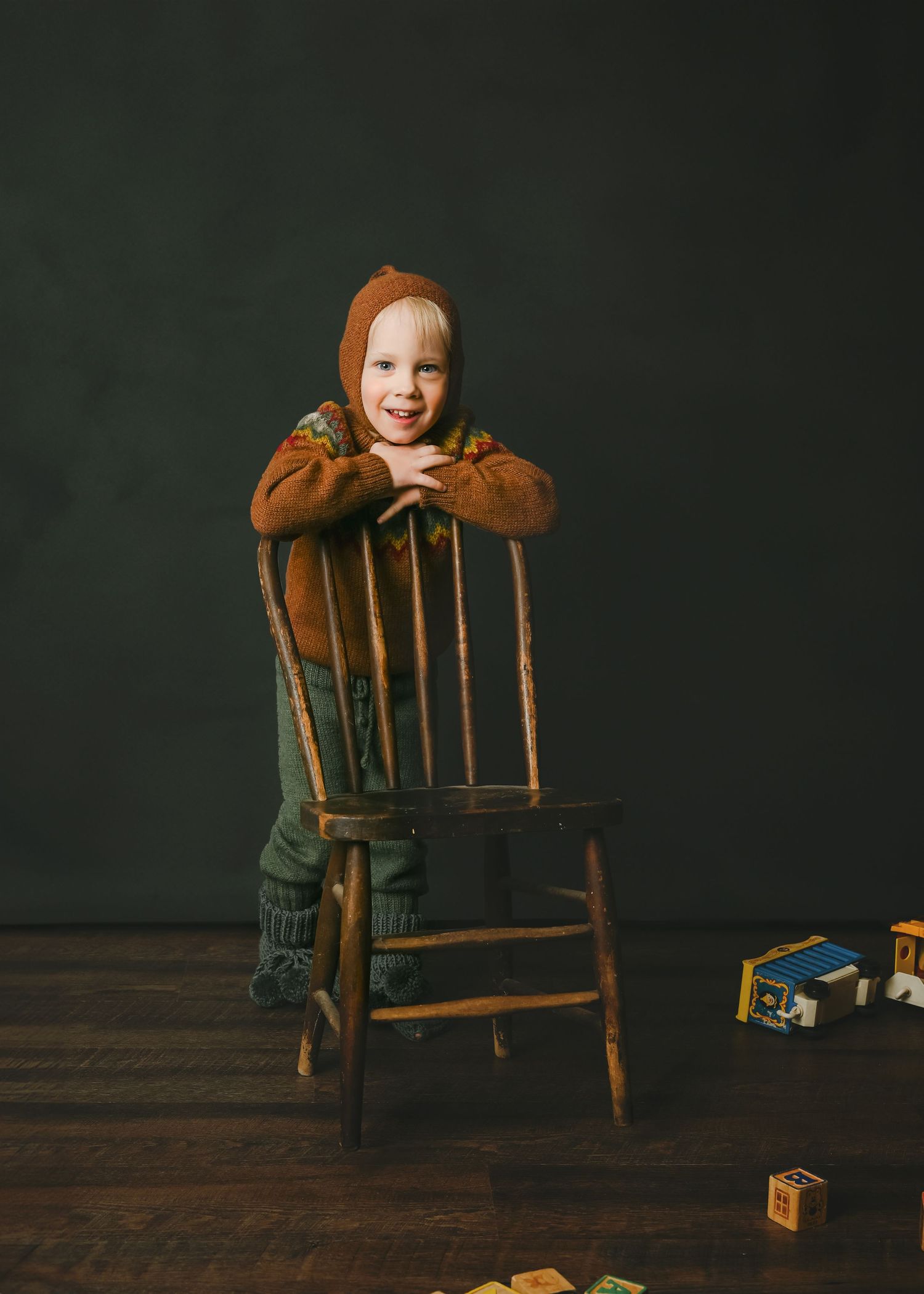 Child in brown sweater leans on wooden chair with blocks scattered on floor.