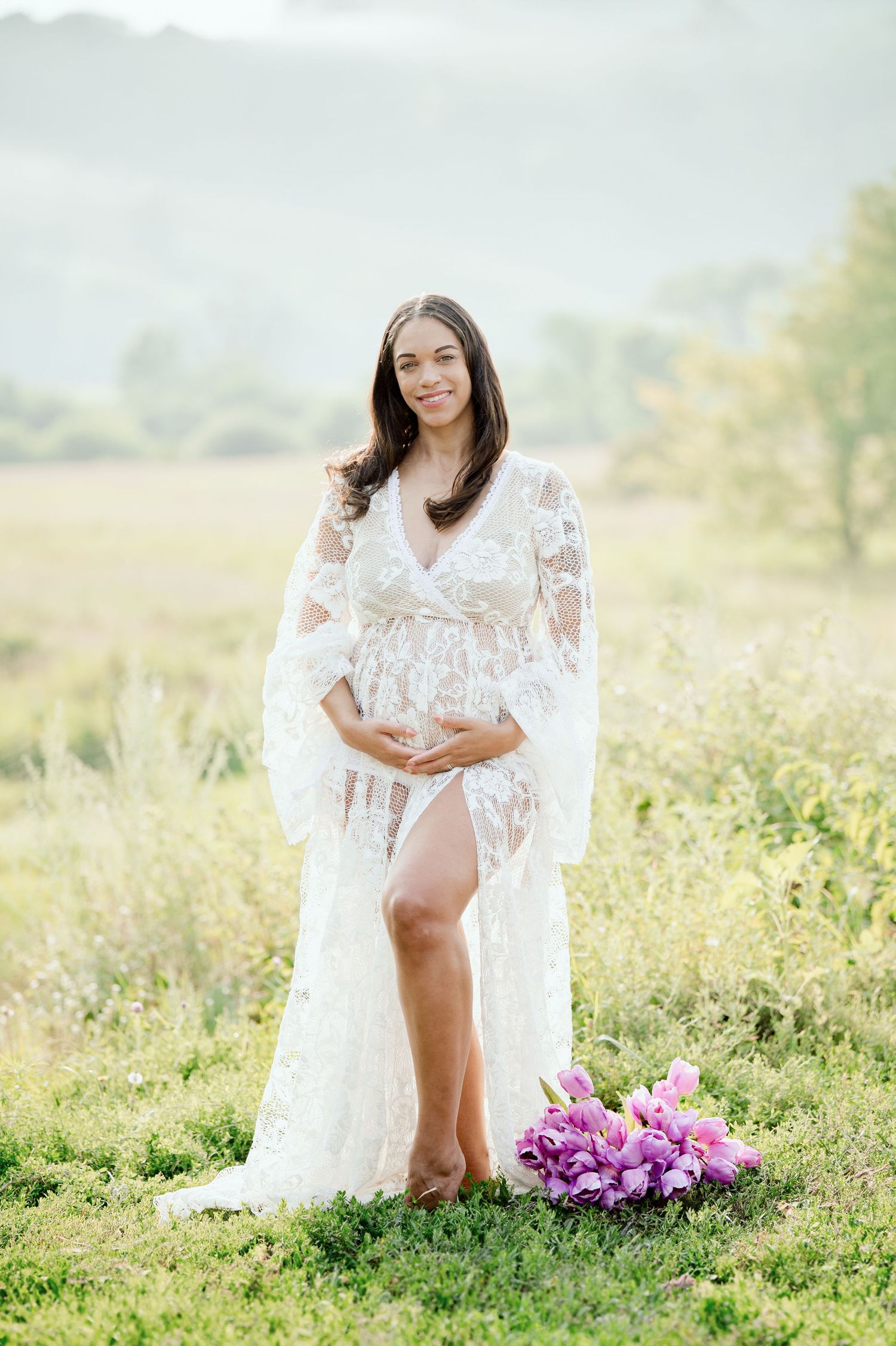 An ethereal white lace maternity gown drapes elegantly in a golden field with purple flowers at sunset.